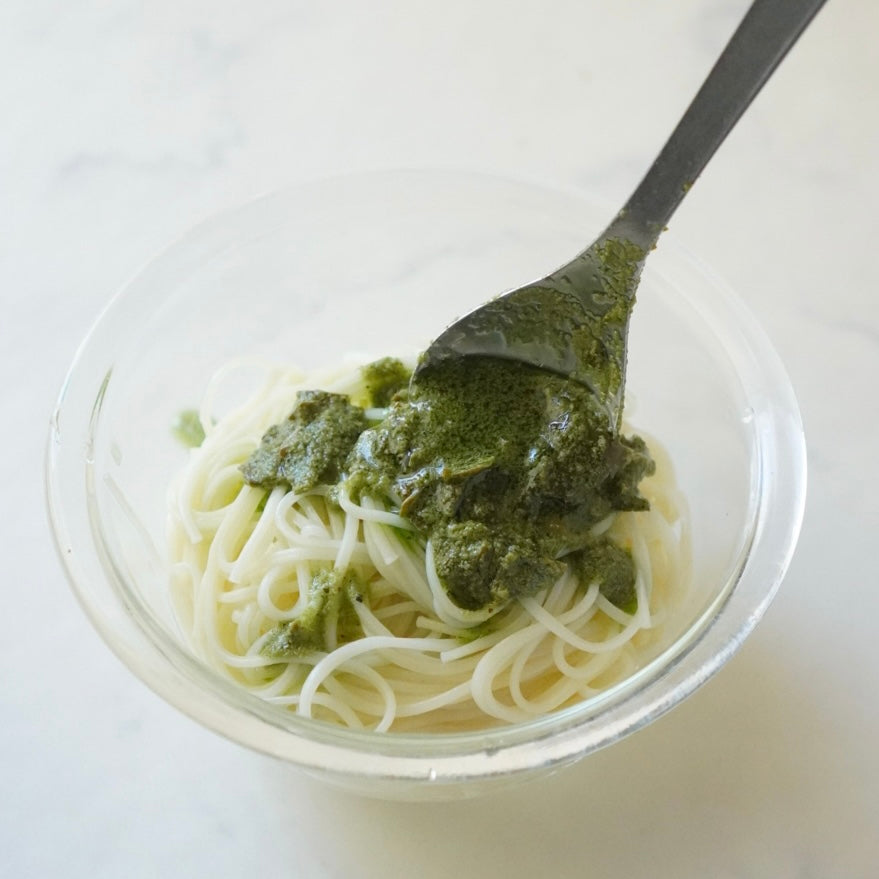 A clear glass bowl of somen noodles with a spoonful of green tea pesto sauce on a light marble surface.