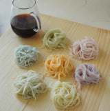 Six small nests of colorful somen noodles in pastel shades are arranged on a light wooden board next to a glass pitcher filled with mentsuyu by yunomi.life